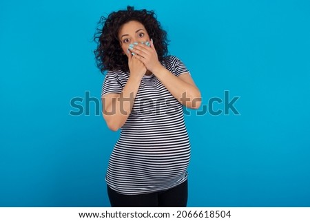 Stunned Young pregnant arab woman wearing striped T-shirt on blue studio background covers both hands on mouth, afraids of something astonishing