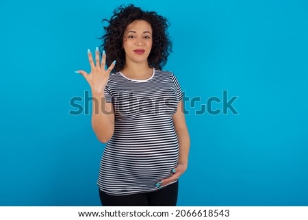 Young pregnant arab woman wearing striped T-shirt on blue studio background smiling and looking friendly, showing number five or fifth with hand forward, counting down