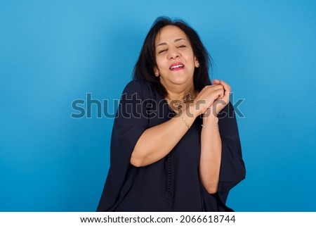 Dreamy middle aged Arab woman standing against blue background with pleasant expression, closes eyes, keeps hands crossed near face, thinks about something pleasant