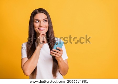 Photo portrait of thoughtful curious woman smiling touching chin keeping mobile phone isolated on bright yellow color background