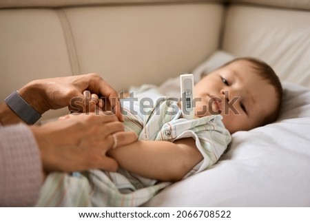 Worried young mother sitting on sofa beside her sick son with high fever. Mom measures temperature using thermometer of sick child lying under blanket at home. Royalty-Free Stock Photo #2066708522