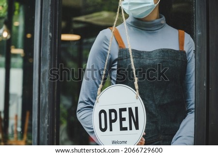 Lockdown. waitress staff woman wearing protection face mask turning OPEN sign board on glass door in modern cafe coffee shop, cafe restaurant, retail store, small business, food and drink concept