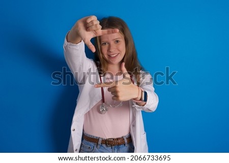 Young caucasian doctor woman wearing medical uniform and stethoscope over blue background making finger frame with hands. Creativity and photography concept.