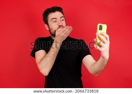 Young caucasian man wearing black t-shirt over red background blows air kiss at camera of smartphone and takes selfie, sends mwah via online call.