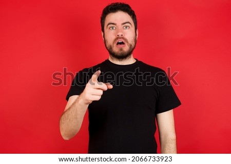 Shocked Young caucasian man wearing black t-shirt over red background points at you with stunned expression