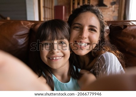 Happy young Latin mum and cute daughter girl taking selfie with face touch, holding smartphone, gadget, looking at camera, smiling. Cheerful mother and kid having fun at home. Close up portrait