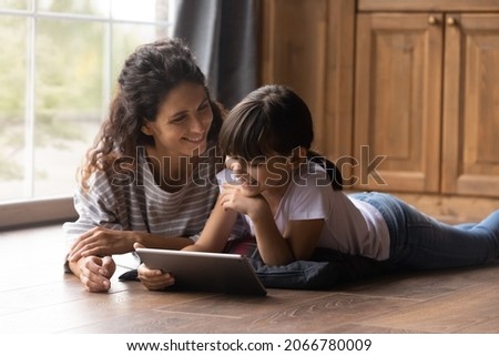 Happy Latin mom and girl resting on heating parquet floor in kitchen, using tablet, laughing, watching movie, making video call, reading ebook on internet together. Mother and daughter at home Royalty-Free Stock Photo #2066780009