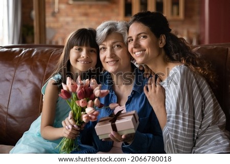 Happy kid girl, mom, grandma celebrating 8 march, birthday, holding bunch of flowers, tulips, gift box, sitting on couch at home, hugging, looking at camera, smiling. Three family generations portrait
