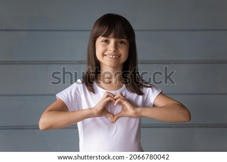 Happy joyful 7s girl making hand heart shape. Cute kid showing love, care sign, looking at camera, smiling. Childhood, childcare, charity, donations, insurance concept. Head shot portrait