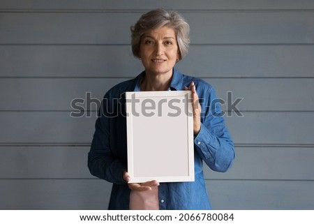Smiling grey haired senior lady head shot. Positive 60s mature adult woman holding blank empty white picture frame, standing at studio background, looking at camera. Female portrait