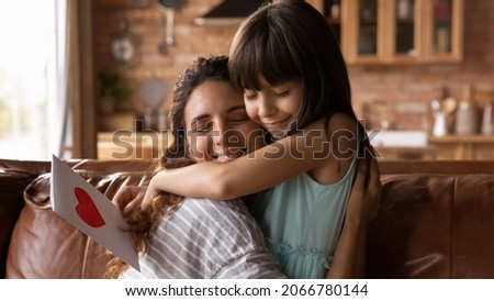 Happy Latin mom and daughter girl celebrating mothers day, 8 march, birthday, hugging on couch at home, receiving gift. Kid giving handmade greeting card to grateful mommy. Motherhood concept