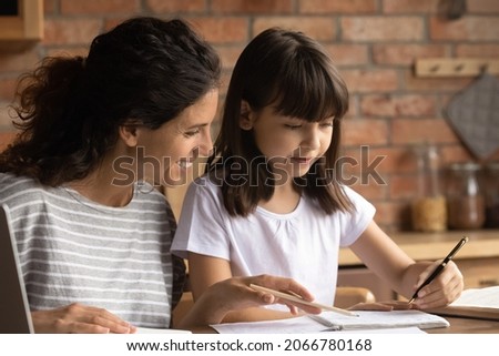Happy mom helping primary schoolkid daughter to do homework. Nanny, sitter, teacher explaining school task to child, teaching kid to write in copybook at home. Homeschooling concept Royalty-Free Stock Photo #2066780168