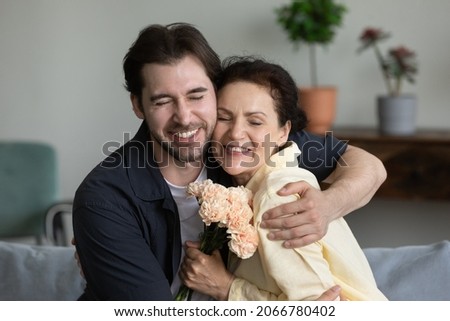 Happy young grown up 30s son cuddling middle aged retired mother, congratulating with birthday or women's day at home, presenting flowers. Bonding two generations family celebrating special occasion. Royalty-Free Stock Photo #2066780402