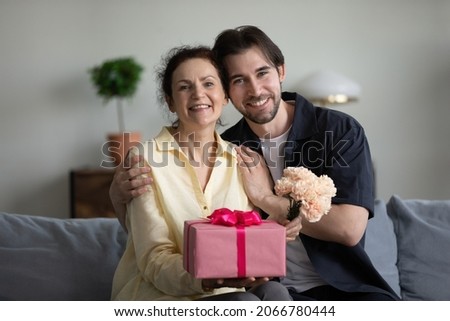 Portrait of bonding young grownup son and sincere joyful mature retired mother sitting on couch, holding bouquet of flowers and wrapped gift in hands, birthday or special occasion celebration. Royalty-Free Stock Photo #2066780444
