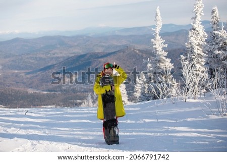 Woman snowboarder in ski clothing poses with a board against a background of blue mountains. Yellow jacket with a hood, knitted hat, ski goggles. Healthy lifestyle. Sports concept. Selective focus.
