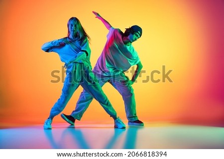 Dancing couple. Stylish man and woman dancing hip-hop in casual youth clothes on gradient yellow red background at dance hall in neon light. Youth culture, hip-hop, fashion, breakdance, action and ad