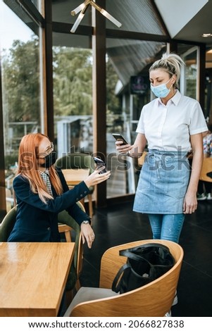 Young attractive business woman sitting in a restaurant or cafe bar and showing digital Covid-19 immunization pass certificate to the waitress. Pandemic and global security measures concept.