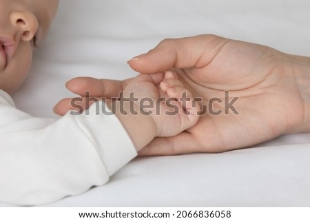 Crop close up loving mother touching small baby hand, sleeping on white bed, family enjoying tender moment, caring young mom holding cute infant newborn arm, motherhood and tenderness concept
