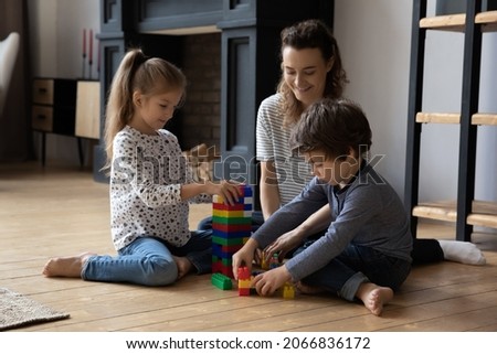 Happy millennial mother or nanny playing toys with cheerful little kids boy girl sitting on warm heated wooden floor in modern living room, constructing building with plastic blocks, playtime activity