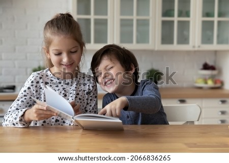 Happy little children siblings reading paper book, enjoying funny story, watching colorful pictures, enjoying spending time together, sitting at table in modern kitchen, entertaining activity concept.