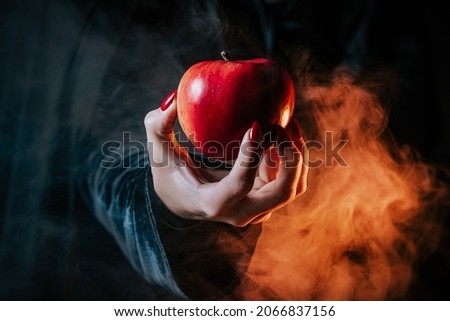 Woman as witch in black offers red apple as symbol of temptation, poison. Fairy tale, white snow wizard concept. Spooky halloween, cosplay. Smoke, haze background. Royalty-Free Stock Photo #2066837156