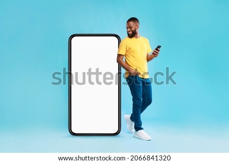 Cheery millennial black guy with mobile device looking at giant smartphone on blue background, mockup for mobile app on white screen. Cellphone website template, cool online store ad Royalty-Free Stock Photo #2066841320