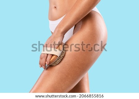 Close up cropped view of young woman making anti cellulite or lymphatic drainage thigh massage. Millennial lady dry brushing her legs, exfoliating skin, taking care of her body, blue studio background