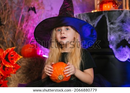 Little cute blonde girl, dressed as a witch on a gloomy background, holds a pumpkin. Decorations for All Saints Day. Halloween costume. Child in a sorceress hat