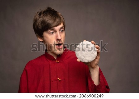 Young handsome tall slim white man with brown hair shocked examining pu erh tea cake in red shirt on grey background