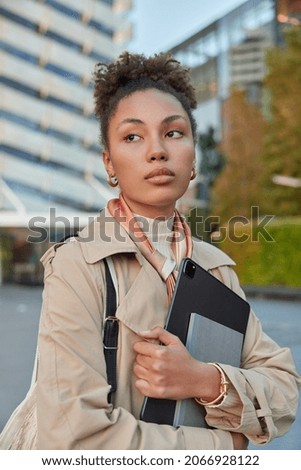 Vertical shot of thoughtful woman dressed in outerwear holds modern tablet and notebook focused away walks outside retrurns from work blurred background. People lifestyle technology concept.