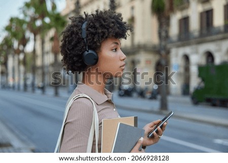 Thoughtful curly female student carries textbooks and notepads holds modern smartphone searches popular songs via cellular media application for listening on leisure stands on empty road in city
