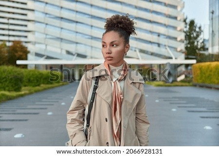 Photo of thoughtful curly haird woman wears beige coat and tied kerchief arouns neck walks outdoors passes by modern city buildings carries bag going to have meeting with someone. Urban lifestyle