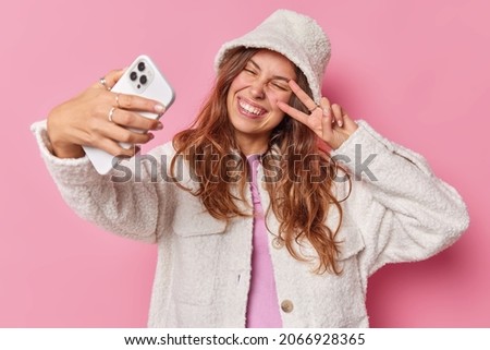 Happy positive young woman makes selfie on smartphone shows peace sign over eye dressed in atificial fur coat and hat enjoys video call isolated over pink background smiles with white teeth.