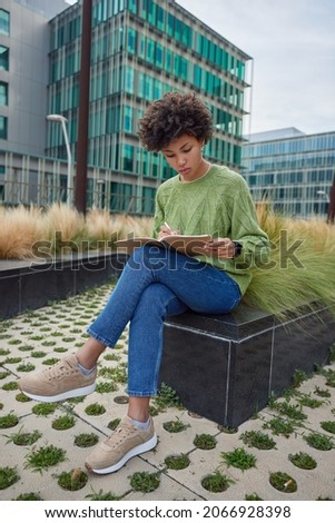 Curly haired female student writes organisation plan in notebook for education uses pen to make notes processes information for university course work wears sweater jeans and sneakers sits outdoors