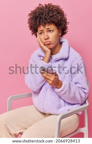 Upset curly haired woman sits on chair uses mobile phone received bad sms feels very unhappy wears fur jacket and trousers isolated over pink background feels offended after quarrel with boyfriend Royalty-Free Stock Photo #2066928413