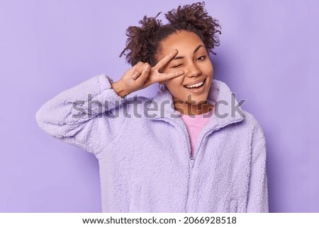 Joyful young woman with hair buns winks eyes makes peace sign over eye smiles happily wears fur coat makes disco finger gesture isolated over purple background. Monochrome shot. Positivity lifestyle