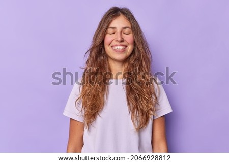 Portrait of attractive teenage girl with long dark hair rouge cheeks and white teeth smiles happily keeps eyes closed makes wish or dreams dressed in casual t shirt isolated over purple background Royalty-Free Stock Photo #2066928812