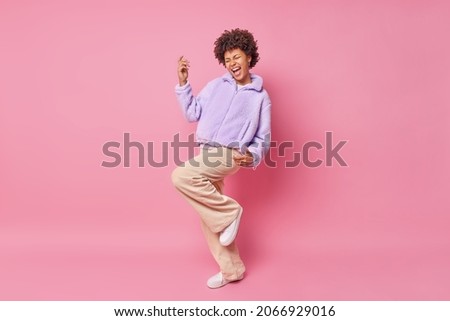 Studio shot of happy curly haired woman makes triumph dance exclaims from joy wears purple fut jacket trousers and white sneakers isolated over pink background with blank space for your promotion