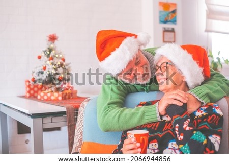 Senior couple in warm clothing and santa hat holding each others hands in front of decorated christmas tree at home. Loving old romantic heterosexual couple celebrating christmas festival together