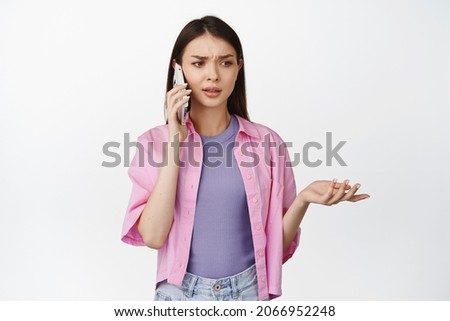 Confused woman talking on mobile phone with puzzled face expression, calling on smartphone and shrugging, standing over white background