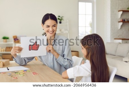 Happy proud female psychologist, therapist or teacher showing thumbs up paper card to little child. Kid receiving positive feedback from English language tutor. Therapy, learning, and success concept