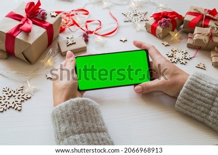 Woman holding smartphone with green screen horizontal position on white wooden table background with Christmas gifts. Mobile Phone with Chroma Key. Close up hands.