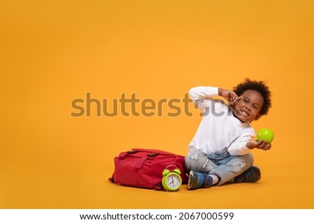 Black child boy 3 years, Student kid holding green apple in his hand and thinking point to head while sitting with school bag and alarm clock. Isolated portrait on yellow background with copy space.