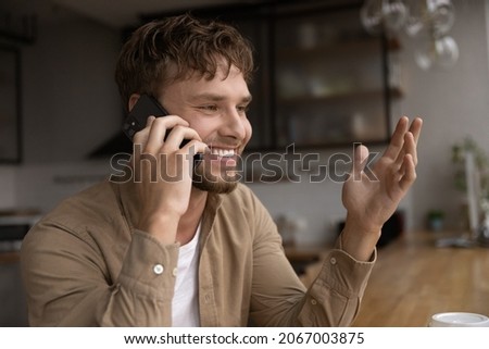 Happy young handsome caucasian man involved in pleasant cellphone call conversation sitting at table, hearing good news, discussing issues calling best friend, communicating distantly at home. Royalty-Free Stock Photo #2067003875