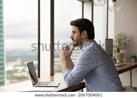 Pensive millennial 30s businessman manager in eyewear looking out of window in distance, thinking on online project problem solution, considering new career challenges, working on computer in office.
