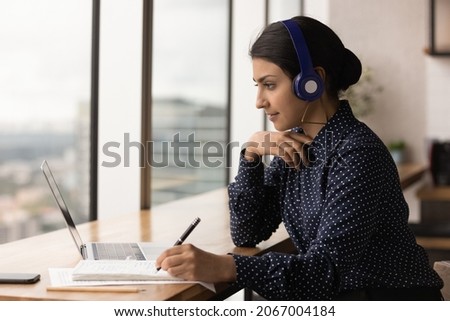 Concentrated young indian woman in headphones looking at computer screen, improving knowledge studying on online courses, writing notes in paper copybook, holding video call preparing for exam. Royalty-Free Stock Photo #2067004184