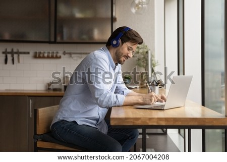 Happy interested millennial generation man in headphones listening educational lecture, studying on online courses in modern kitchen, handwriting notes in paper copybook, improving knowledge. Royalty-Free Stock Photo #2067004208