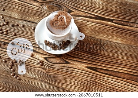 Top view of ceramic cup of hot cappuccino coffee latte with drawing picture of heart shape with cinnamon or cocoa on milk foam, different plastic stencils, coffee beans on wooden table, copy space