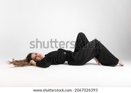 Girl, blonde with curly hair in black pants and a shirt posing while sitting on a white background barefoot.