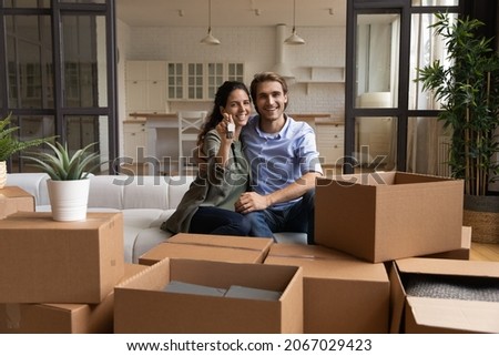 Happy couple enjoy relocation day, sit on sofa smile look at camera showing bunch of keys from new house, cardboard boxes with belongings nearby. Bank loan, tenant, homeowners family portrait concept Royalty-Free Stock Photo #2067029423